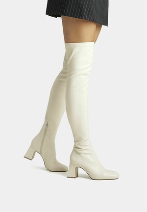 Jambes portant des bottes à talons écru au-dessus du genou avec des fermetures éclair latérales et une jupe noire à fines rayures sur un fond clair.