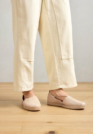 Person iført beige vidde bukser og lyserøde espadrille-sko med ankelstropper står på trægulv.