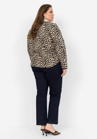 Jachetă cu imprimeu de leopard pe un fundal bej, cu contur negru și mâneci lungi, asortată cu pantaloni de culoare albastru închis și sandale cu toc cu imprimeu de leopard.