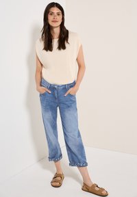 Beige T-shirt met korte mouwen, lichtblauwe cropped jeans met opgerolde manchetten en bruine sandalen. De outfit heeft een casual, relaxte pasvorm.