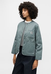 Γυναίκα με σγουρά μαλλιά που φοράει γαλάζιο μπουφάν με καπιτονέ λεπτομέρειες πάνω από μια ασπρόμαυρη ριγωτή μπλούζα και σκούρο τζιν με φαρδιά μπατζάκια, κοιτάζοντας προς τα αριστερά.