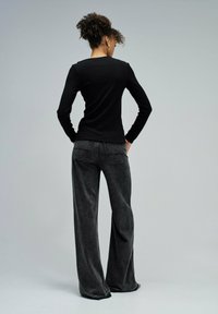 Une femme aux cheveux bouclés se tient de dos, portant un haut noir à manches longues et un jean gris foncé ample, contre un fond gris uni.