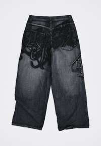 Μαύρα και γκρι ξεθωριασμένα τζιν με φαρδιά μπατζάκια, διακοσμημένα με μαύρη κεντητή λεπτομέρεια στις τσέπες και ραμμένο μπαλώμα με αριθμό στο δεξί μηρό.