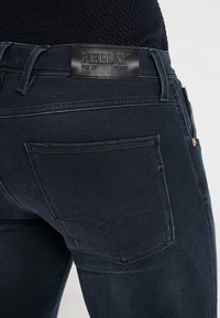 Mörkblå denimjeans med en figursydd design, med en läderetikett vid midjan och ett subtilt stickmönster på bakfickorna.