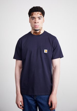 T-shirt en coton bleu marine à manches courtes, avec une poche poitrine et un patch logo prominent, associé à un jean en denim. Design simple et minimal.