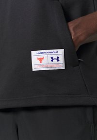 Felpa nera con etichetta Under Armour, logo rosso del toro e logo blu, texture morbida e design classico.