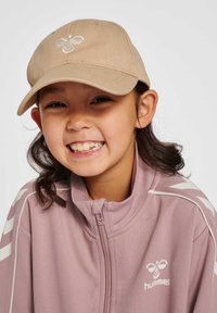 En tanfärgad keps med broderad logotyp, som bärs av ett barn i en rosa zip-up jacka med vita ränder och logotyp, leende med synliga tänder.