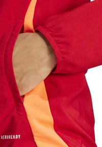 Veste rouge avec une doublure intérieure orange, dotée de poignets élastiques et d'une poche. Le matériau semble lisse et léger, adapté pour les vêtements de sport.