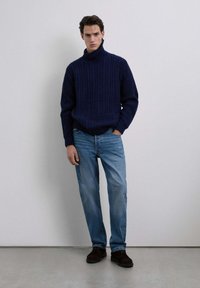 Un pull-over col roulé en tricot torsadé bleu marine associé à un jean bleu clair et des chaussures marron foncé, créant un look décontracté et texturé.