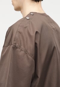 Homme portant une chemise marron avec un motif en grille, comportant deux grands boutons sur la couture de l'épaule.
