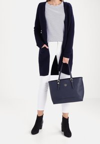 Donna che indossa un cardigan blu scuro, una maglietta a righe, pantaloni bianchi e stivaletti neri alla caviglia, mentre tiene una borsa blu scuro per i manici.