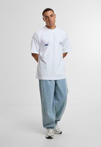 Camiseta de algodón blanca con un corte holgado, mangas cortas y detalle de logo azul en el pecho, combinada con unos jeans de mezclilla azul claros y anchos, y zapatillas plateadas.