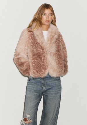 Jeune femme aux longs cheveux blonds portant une veste duveteuse rose et marron et un jean bleu déchiré sur un fond uni clair.