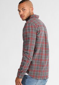 Camisa de cuadros en rojo y gris, con botones, hecha de franela de algodón, que presenta mangas largas y un cuello clásico con un acabado ligeramente texturizado.
