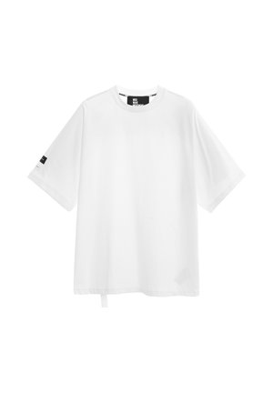 Joven de pie con las manos detrás de la espalda, vistiendo una camiseta blanca de gran tamaño, pantalones cortos negros hasta la rodilla con cadenas, calcetines negros y zapatos negros.