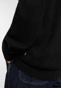 Schwarzer Strickpullover mit geripptem Saum und Bündchen. Das Material scheint weich mit einer strukturierten Oberfläche zu sein. Kleines Logo-Etikett ist an der Seite sichtbar.