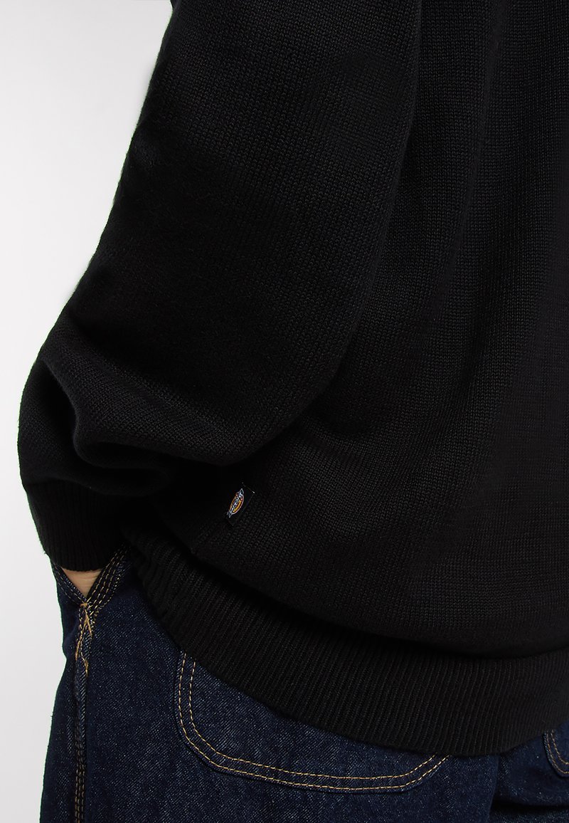 Sort striktrøje med ribbet kant og manchetter. Materialet ser blødt ud med en tekstureret overflade. Lille logoetiket synlig på siden.