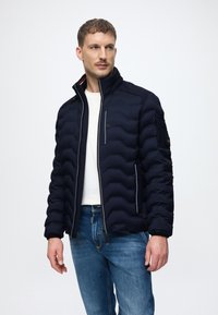 Μπλέ puffer μπουφάν με πλισέ υφή, ψηλό γιακά, μπροστινό φερμουάρ, πλευρικές τσέπες και αντίθετες λεπτομέρειες. Φορεμένο με ένα λευκό πουκάμισο και τζην.