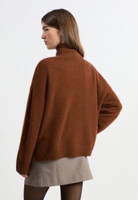 Rdzawy, oversize sweter z wysokim kołnierzem i żebrowanym dołem. Materiał wydaje się miękki i teksturowany. Noszony z szaro wzorzystą spódnicą.