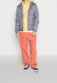 Person med en grå jakke over en gul hoodie, orange bukser og blå og grønne sneakers, der står mod en ensfarvet væg.