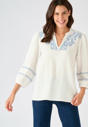 Femme souriante, portant un chemisier blanc texturé avec une broderie florale bleue sur le col et les manches, associé à un jean bleu foncé.