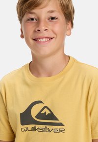 Žluté bavlněné tričko s velkým černým logem Quiksilver na přední straně. Tričko má kulatý výstřih a krátké rukávy.