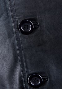 Schwarzer Lederstoff mit sichtbarer Naht. Zwei große schwarze Knöpfe, von denen einer teilweise geschlossen ist, mit einem Vierlochkraftdesign und glänzender Oberfläche.