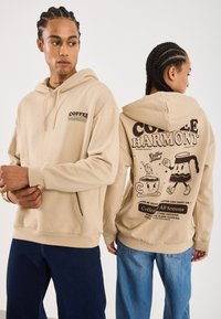 Deux personnes avec des cheveux tressés portent des sweatshirts beige ; le logo à l'avant indique "Coffee Harmony", l'arrière présente des graphiques de tasse et de cafetière avec du texte.