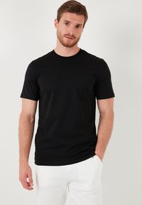 Černé tričko s krátkým rukávem a kulatým výstřihem, vyrobené z měkké látky. Má slim fit střih a hladkou texturu, nošeno s bílými kalhotami.