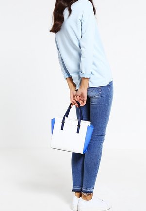 Bolso blanco y azul con forma estructurada, acentos laterales azules y asas cortas negras. Lleva puesto un top azul claro y unos jeans de mezclilla.