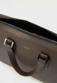 Sac en cuir marron foncé avec une finition texturée, doté de deux poignées, d'une fermeture éclair et d'un logo à l'avant. L'intérieur est en tissu noir.