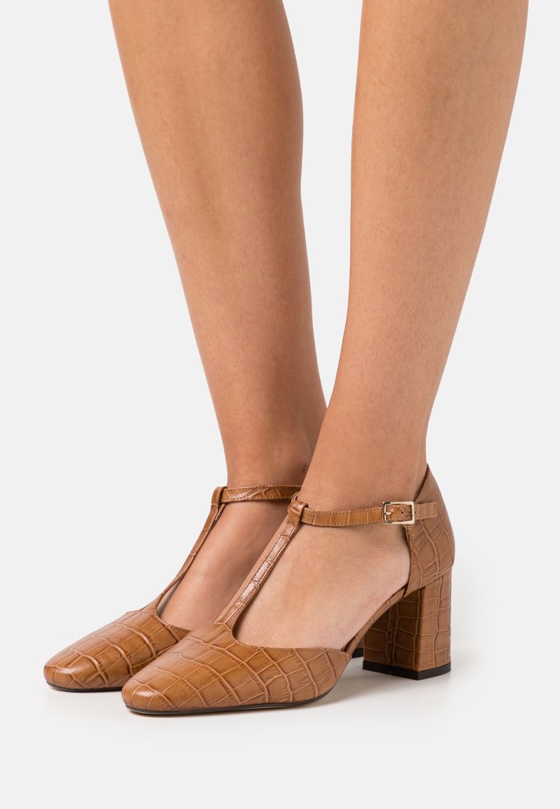 Chaussures à talon bloc en cuir marron à motif crocodile avec bride en T, portées pieds nus sur un fond clair uni.