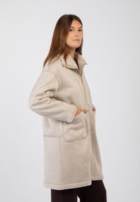 Cappotto in fleece beige con colletto alto, chiusura con zip, tasche frontali e bordi e polsini in morbido cremoso a contrasto.