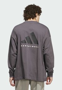 Maglietta grigia a maniche lunghe con scollo rotondo. Presenta un grande logo triangolare nero e il testo "BASKETBALL" sul retro. Materiale in cotone.