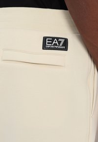 Bézs sportos nadrág sima textúrával, rugalmas derékkal és hátsó zsebében egy fekete EA7 Emporio Armani logó címkével.