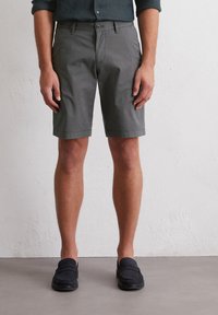 Shorts en coton gris avec un devant plat et des poches latérales, dotés d'une fermeture à bouton, accompagnés de mocassins noirs.