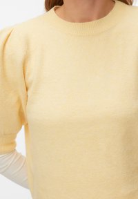 Femme portant un haut en maille jaune pâle à manches courtes bouffantes par-dessus une chemise blanche à manches longues, sur fond blanc.