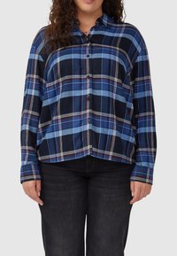 Woman wearing a blue, black, and white plaid button-up shirt with long sleeves paired with black jeans, standing against a plain background.