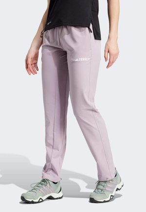 Lilla Adidas Terrex bukser båret af en person med grønne og lavendel atletik sko og sort top, stående på en lys baggrund.