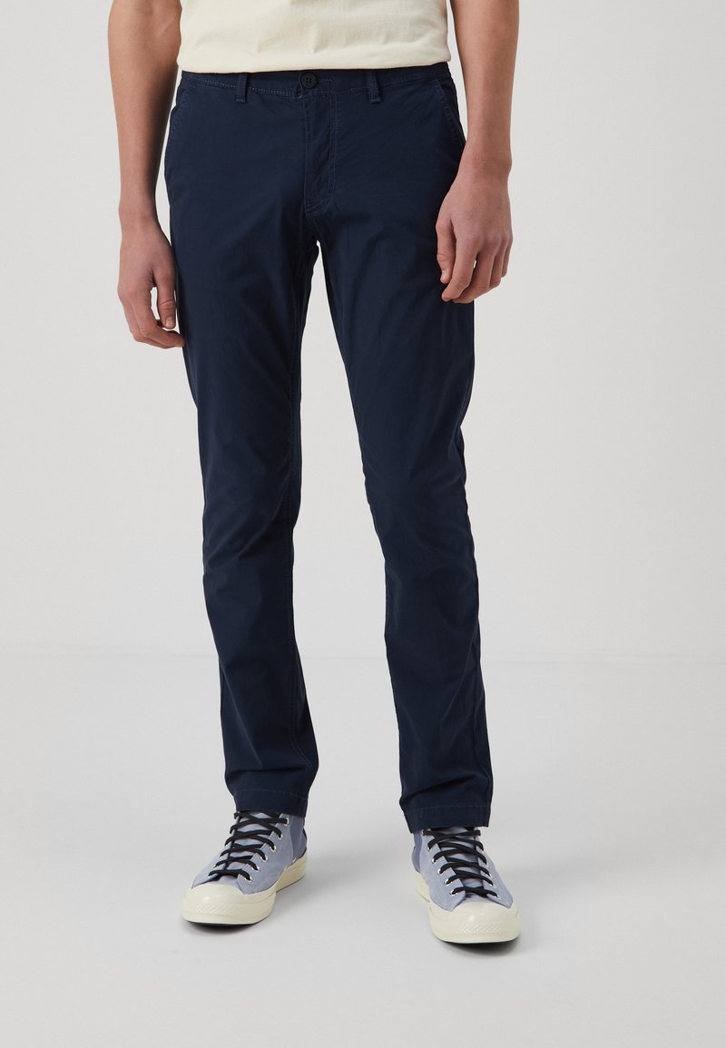 Navy blue cotton twill pants with a slim fit, featuring a button closure and subtle pocket detailing, paired with light blue sneakers.