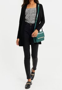 Svart cardigan bärs över en svart och vit mönstrad topp, ihop med svarta skinny jeans och en teal-färgad väska med en texturerad yta.