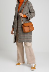 Žena v kostkovaném kabátu, s oranžovým rolákem, béžovými kalhotami s širokými nohavicemi, hořčicovými teniskami a nosící hnědou koženou crossbody kabelku s detailem třásní.