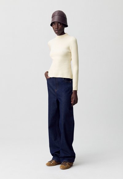 Femme portant un haut à manches longues côtelé crème, un jean large bleu foncé, un chapeau en cuir marron et des chaussures lacées beige sur un fond uni.