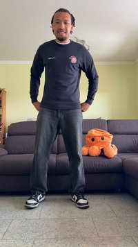 Fekete, hosszú ujjú póló logóval, szürke farmernadrág és fekete sportcipő fehér részletekkel. Plüss narancssárga polip játék egy sötét kanapén a háttérben.