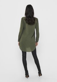 Tunique vert olive à manches longues en tissu léger, avec un ourlet arrondi. Portée avec des leggings noirs et des bottines à talons.