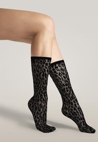 Ben klædt i gennemsigtige, sorte knæstrømper med leopardmønster mod en ensfarvet, lys baggrund.