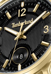 Zlato in črna analogna ura z zrnatim črnim številčnico, bold oznakami ur in kazalci s svetlečimi poudarki. Na voljo blagovna znamka "Timberland".