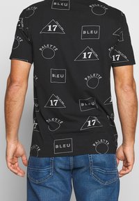 Camiseta negra con patrones geométricos y texto en blanco. Presenta triángulos, círculos y cuadrados. El material parece suave y tiene un ajuste regular.