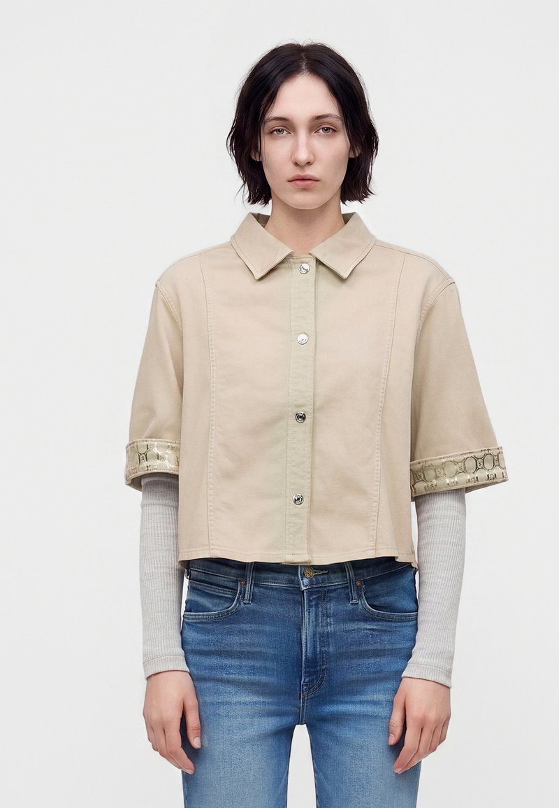 Personne aux cheveux courts et foncés portant une chemise beige à manches courtes boutonnée par-dessus un haut gris clair à manches longues et un jean bleu sur un fond uni.