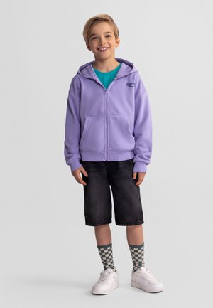 Uśmiechnięte dziecko stoi, ubrane w fioletową bluzę z zamkiem, turkusową koszulkę, czarne krótkie spodenki do kolan, kraciaste skarpetki i białe trampki na szarym tle.